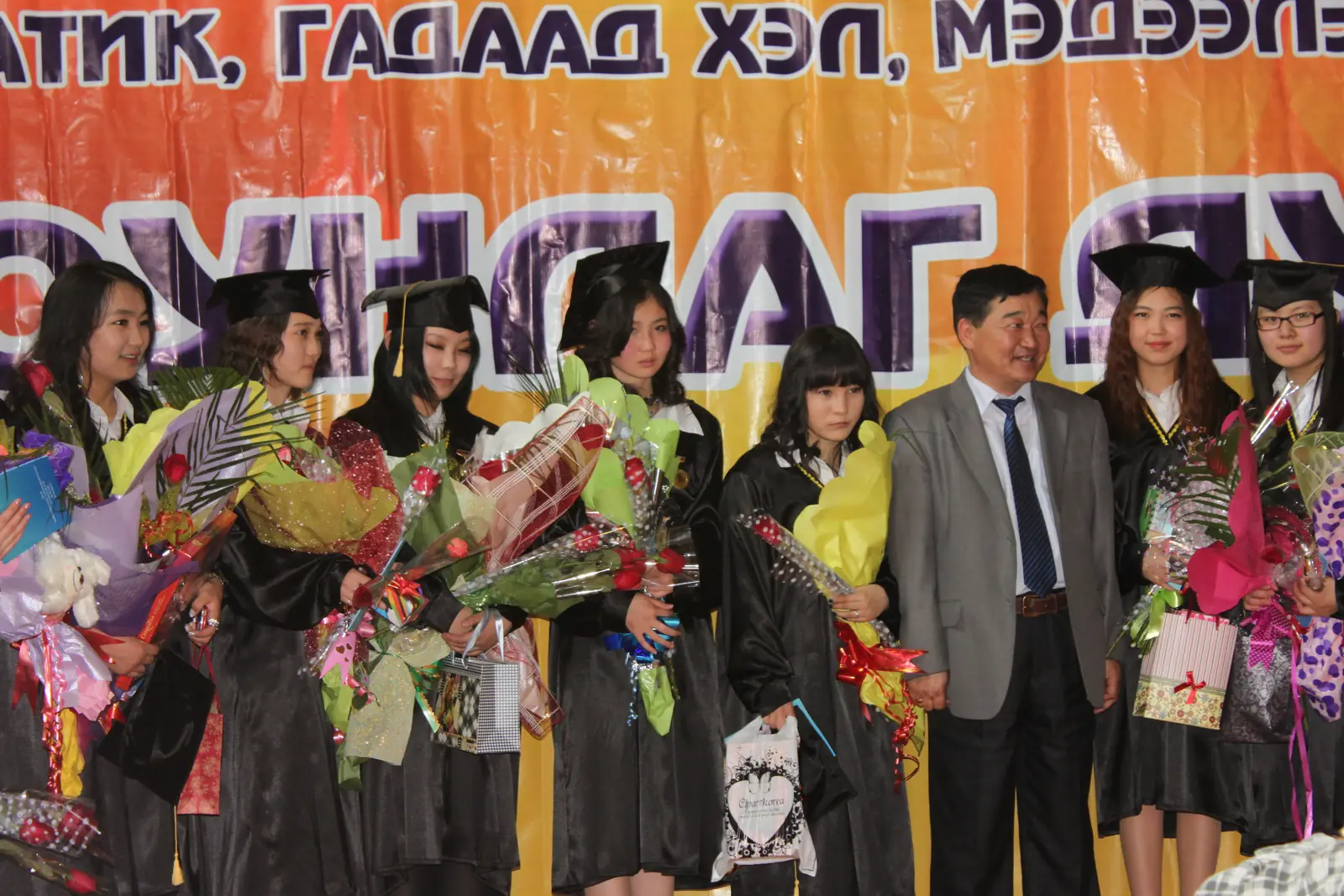 头戴学位帽、身穿学位服的毕业生手捧鲜花，与老师一同站在台上