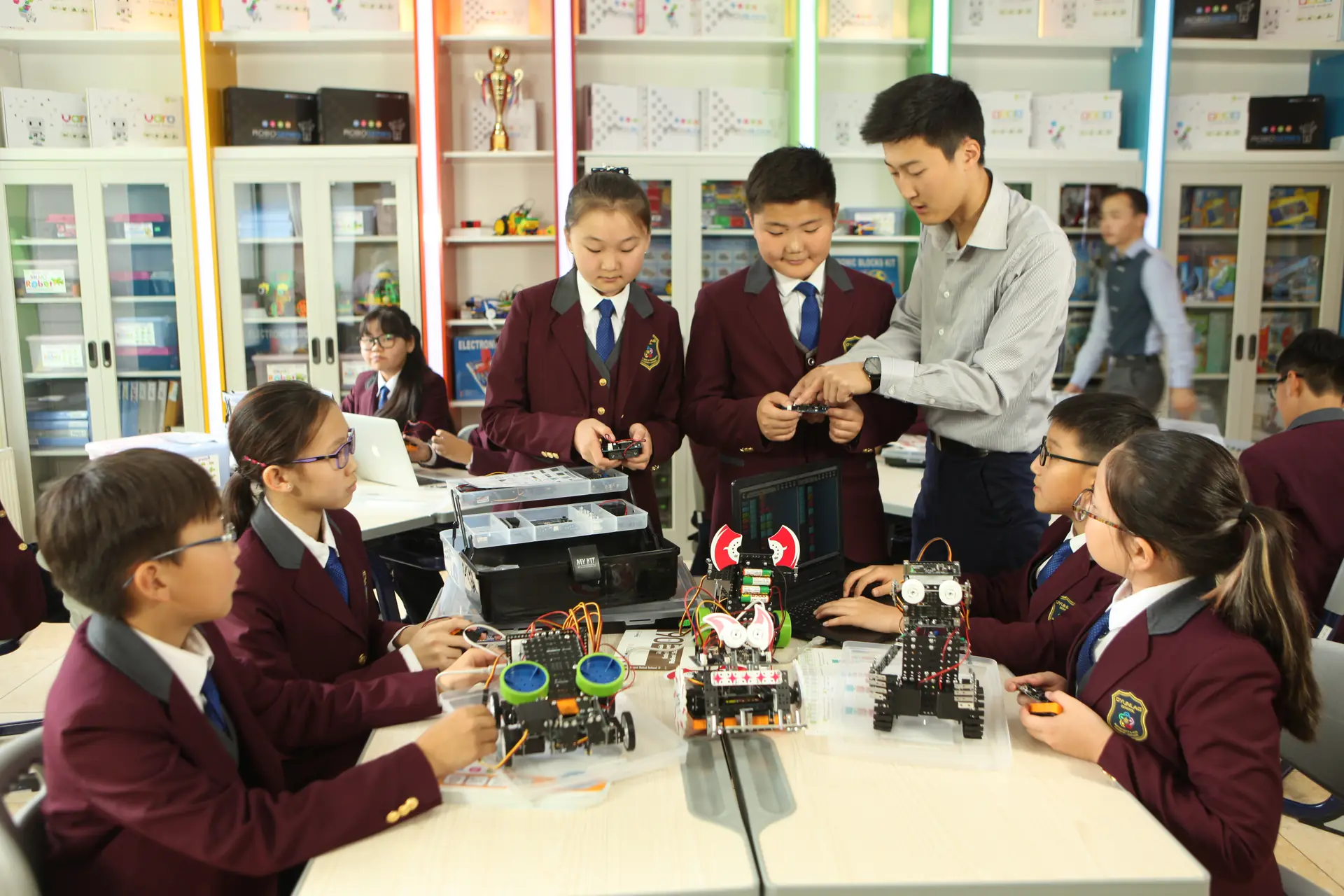 身穿酒红色西装外套的学生和老师在机器人实验室里搭建并测试小型机器人